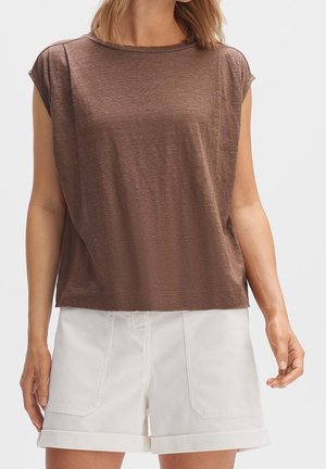 Vrouw draagt een mouwloos bruin topje en witte korte broek met omslag, staat tegen een effen lichte achtergrond.