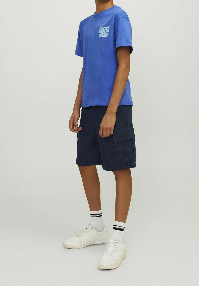 Personne portant un t-shirt bleu "Frozen Dreams", un short cargo bleu marine, des chaussettes blanches avec des rayures noires, et des baskets blanches, se tenant de profil.