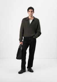 Σκούρο πράσινο πλεκτό πουλόβερ με γιακά και φερμουάρ, συνδυασμένο με μαύρο παντελόνι. Μαύρα παπούτσια και δομημένη μαύρη τσάντα tote. Απλό φόντο.