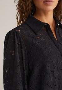 Črna čipkastna majica s ovratnikom, ki ima cvetlični vzorec, z gumbnim zapiranjem in teksturiranimi rokavi. Okrščena s subtilnim sijajem.