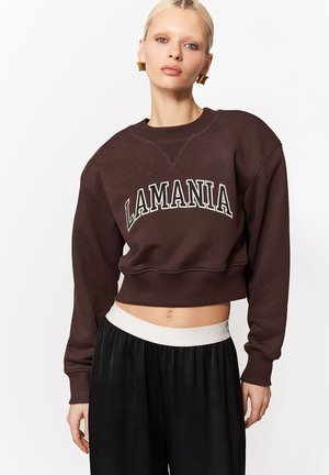 Blondynka w brązowej krótkiej bluzie z napisem "LAMANIA" i czarnych spodniach z białym pasem, stojąca na białym tle.