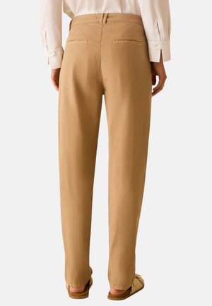 Frau trägt beige gerade geschnittene Hose, weißes langärmeliges Hemd und beige Slipper, von hinten vor einfachem Hintergrund gezeigt.