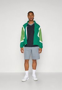 Grønn og hvit jakke med elastiske mansjetter, svart sportsskjorte, grå shorts og hvite joggesko. Avslappet atletisk antrekk for varmt vær.