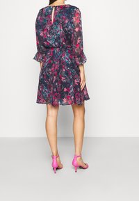 Kvinna klädd i en marinblå blommig klänning med rosa och teal-accenter, 3/4-ärmar, i kombination med klara rosa högklackade sandaler, stående vänd bort.