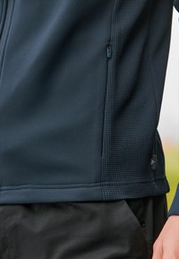 Chaqueta deportiva en color navy hecha de una tela suave con paneles laterales texturizados, que cuenta con un bolsillo con cremallera y un sutil detalle del logo.