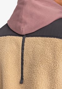 Fleecejacke mit beigem, strukturiertem Körper, dunkelgrauem oberen Abschnitt und einer pinken Kapuze. Merkmale sind eine Mittelrückenaht mit strukturiertem Detail.