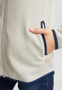 Vilnas jaka krēmkrāsā ar tumši ziliem akcentiem. Aprīkota ar rāvējslēdzēju priekšpusē, sānu kabatām un koka etiķetes detaļu. Gluda tekstūra un brīvs piegriezums.