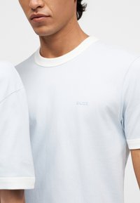 Άνδρας που φοράει λευκό T-shirt με κοντά μανίκια και μικρό κεντητό λογότυπο "BOSS" στο στήθος, με μονόχρωμο φόντο.
