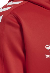 Roter sportlicher Kapuzenpullover mit weißen Chevron-Akzenten auf den Schultern. Hergestellt aus glattem Stoff, mit einem weißen Logo auf der Brust und einer strukturierten Oberfläche.