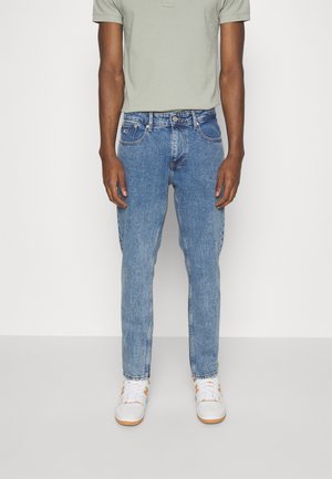 Uomo con polo a maniche corte grigio chiaro, jeans blu a gamba dritta e sneakers bianche in piedi davanti a uno sfondo bianco neutro.