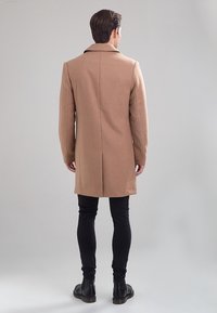 Homme portant un manteau camel jusqu'aux genoux, un pantalon noir et des bottes noires, se tenant de dos contre un fond clair uniforme.