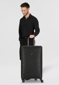 Valigia rigida nera con motivo in rilievo, angoli arrotondati, doppie ruote girevoli e manico estensibile. Logo visibile sulla parte anteriore.