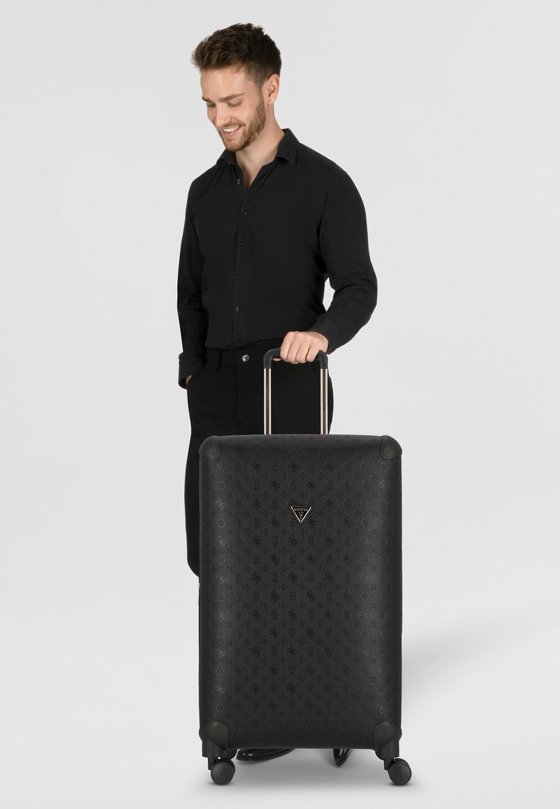 Valigia rigida nera con motivo in rilievo, angoli arrotondati, doppie ruote girevoli e manico estensibile. Logo visibile sulla parte anteriore.