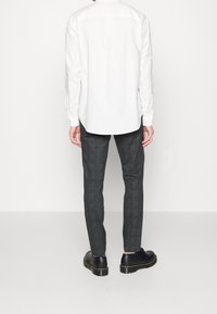 Chemise blanche à manches longues avec ourlet arrondi, associée à un pantalon à carreaux gris foncé et des chaussures noires à semelle épaisse avec des coutures jaunes.