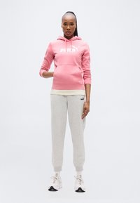 Ροζ φούτερ με τσέπη καγκουρό και λευκό λογότυπο, συνδυασμένο με γκρι φόρμες. Το μοντέλο φοράει λευκά και γκρι αθλητικά παπούτσια. Λείο ύφασμα.