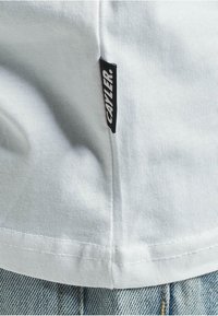 Weißes T-Shirt aus strukturiertem Stoff, mit einem schwarzen Etikett, auf dem „CAYLER“ an der Seitennaht steht. Kombiniert mit Denim, mit einem gesteppten Saum.