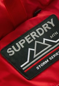Sarkana auduma ar melnu plecu, uz kura balti burti ir uzrakstīts "SUPERDRY", kalnu grafika un balti burti apakšā "STORM SERIES".