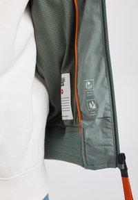 Vihreä tuulenpitävä ja vedenpitävä takki, jonka sisäpuolella on verkkovuori, oranssit yksityiskohdat ja etiketti, joka näyttää hoito-ohjeet ja ominaisuudet.