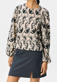 Femme portant un blouse longue beige à manches longues avec des broderies florales noires et une mini-jupe noire avec une fente à l'avant.