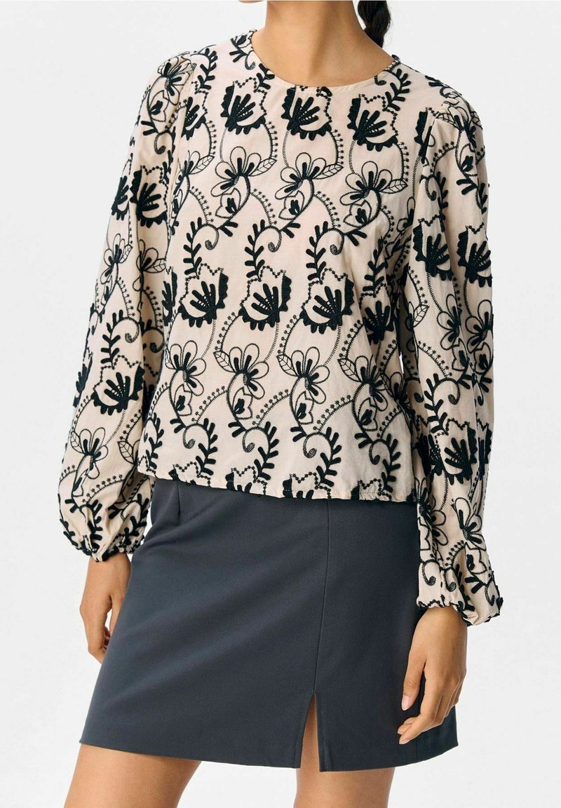 Femme portant un blouse longue beige à manches longues avec des broderies florales noires et une mini-jupe noire avec une fente à l'avant.