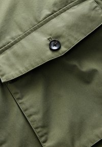 Olive green fabric with a smooth texture, featuring a flap pocket and a black button detail. The stitching is neat and consistent.