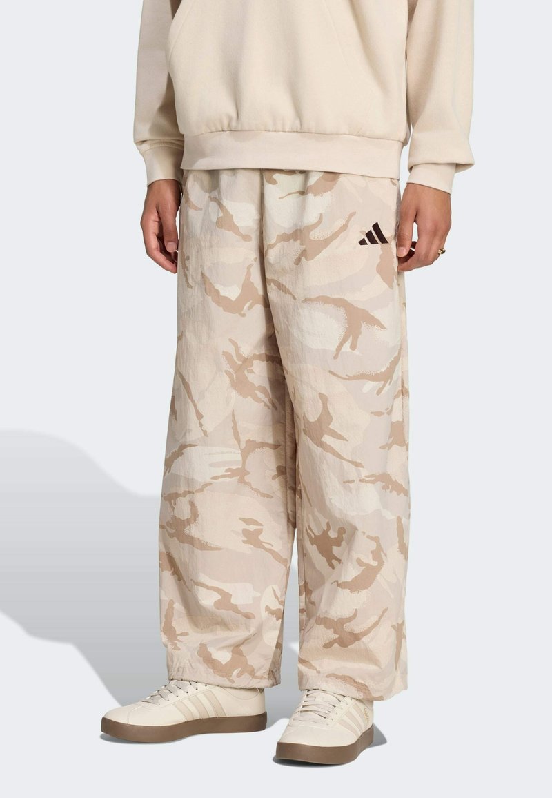 Personne portant un pantalon large camouflage crème et beige, un sweat assorti et des baskets écrues avec des semelles marron.