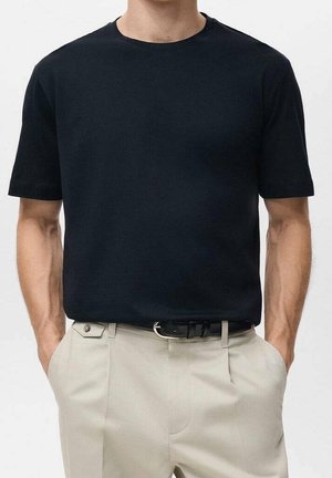 Homme portant un t-shirt noir uni à manches courtes, rentré dans un pantalon beige avec une ceinture noire, mains dans les poches.