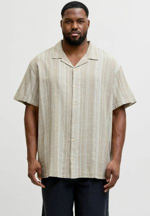 Mann stående iført en beige kortermet skjorte med hvite vertikale striper og svarte bukser mot en ensfarget bakgrunn.