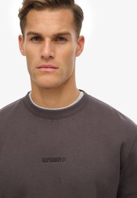 Szara bluza z ribbed crew neck, z haftowanym logo "SUPERDRY" na środku. Materiał wydaje się być miękki z teksturowanym wykończeniem.