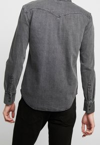 Harmaa farkkupaita, jossa on pitkät hihat, napit hihansuissa, takana kärkipäinen yläosa ja hieman kaartuva helma. Kankaan pinta on teksturoitu.