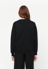 Mujer con cabello largo castaño que lleva un sudadera negra lisa y pantalones negros, mostrada de espaldas contra un fondo blanco.
