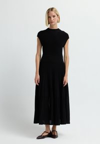 Abito nero a costine con collo alto e maniche corte, caratterizzato da una gonna plissettata. Indossato con scarpe basse scure.