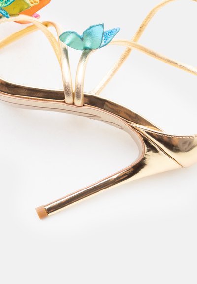 Χρυσό πέδιλο με ψηλό τακούνι με λεπτά λουράκια και μια μπλε διακοσμητική πεταλούδα κοντά στο λουράκι του αστραγάλου, σε λευκό φόντο.