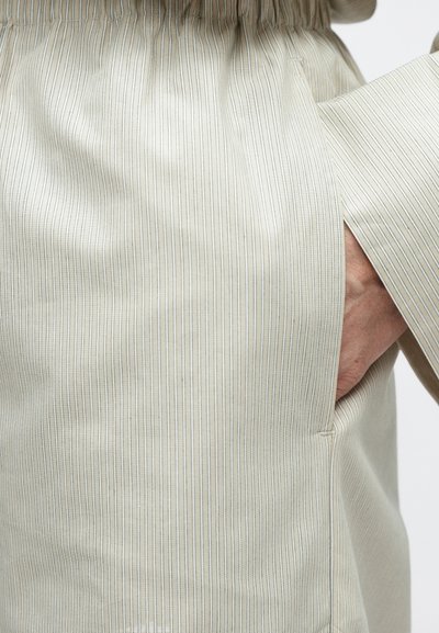 Világos krémszínű anyag vékony, függőleges csíkokkal, oldalhasítékokkal és látható zsebbel. Simább textúrájú és rugalmas derékrésszel.