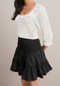Vrouw in een witte gehaakte lang-sleeve top met bloemendetail en een zwart hoge taille gelaagd rokje voor een effen muur.