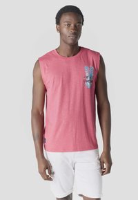 Giovane uomo che indossa una maglietta rosa senza maniche con stampa di palme e pantaloncini bianchi strappati, su uno sfondo grigio uniforme.