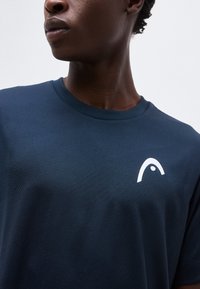 Κοντινό πλάνο ενός ατόμου που φοράει μπλε ναυτικό μπλουζάκι με στρογγυλή λαιμόκοψη και ένα μικρό λευκό στυλιζαρισμένο λογότυπο στο αριστερό στήθος.