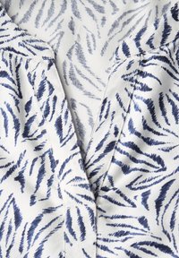 Tessuto bianco con motivo a zebra in rilievo blu navy; presenta un colletto a scollatura a V e una patta con bottoni per la chiusura.
