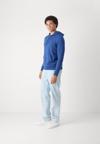 Felpa in cotone blu con tasca a marsupio e cappuccio regolabile, abbinata a jeans di denim chiaro e sneakers bianche. Il modello è in piedi su uno sfondo neutro.