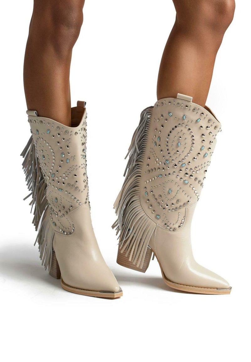 Botas de cowboy de cuero beige con detalles de flecos, adornadas con rhinestones y punta afilada, con un tacón cuadrado y acabado suave.