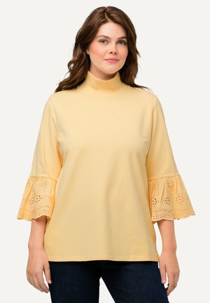 Haut jaune à manches longues avec un col montant et des manches évasées présentant des détails en œillets aux poignets, assorti à un pantalon en denim foncé.