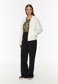 Weißer, strukturiert Bomberjacke, olivfarbenes Grafik-T-Shirt, schwarze weite Hose und schwarze Sneaker mit dicker Sohle.