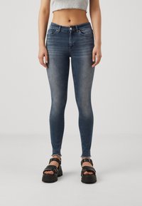 Jeans ajustados de denim azul oscuro, con una cintura de altura media, cinco bolsillos y un ligero desgaste en el dobladillo. Llevados con sandalias negras gruesas.