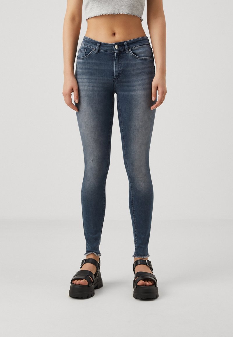 Jeans ajustados de denim azul oscuro, con una cintura de altura media, cinco bolsillos y un ligero desgaste en el dobladillo. Llevados con sandalias negras gruesas.
