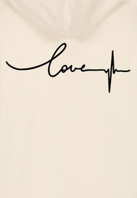 Krem dukserica s kapuljačom koja ima veliki crni kurzivni natpis reči "ljubav" integriran s dizajnom linije otkucaja srca.