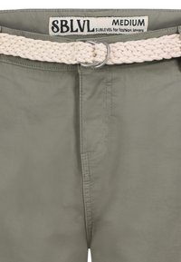 Khaki groene broek met een gladde textuur, voorzien van een geweven crèmekleurige riem en een zilveren metalen gesp. Het label leest "SBLVL MEDIUM."