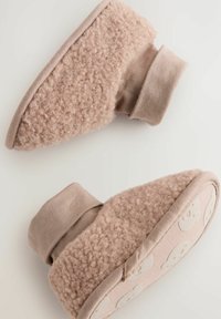 Botines suaves en beige con un upper de peluche, un puño de tela lisa y una suela con un patrón de caras de oso. Forma redondeada con acentos de costura.