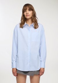 Camicia button-up a righe blu chiaro e bianche con una vestibilità ampia, taschino sul petto e maniche lunghe. Tessuto liscio e a trama intrecciata.