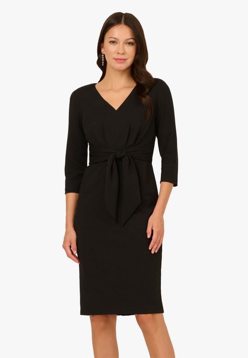 Schwarzes knielanges Kleid aus glattem Material. Es hat einen V-Ausschnitt, Dreiviertelärmel und eine Bindung an der Taille.