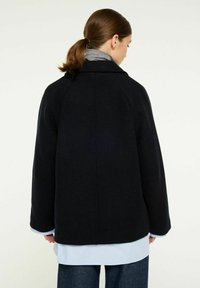 Svart ullkappa med en strukturerad design, raglansärmar och en något översized passform. Har en hög krage och en kontrasterande ljusblå skjorta under.